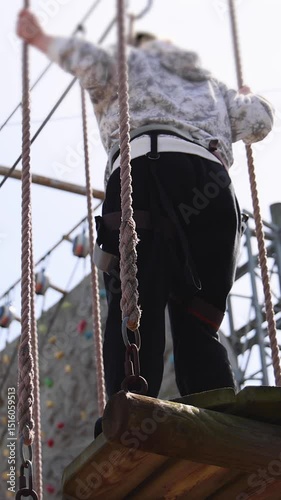 View of high ropes course, kids of climbing in amusement acitivity rope park, passing obstacles and zip line on heights, children teenagers in equipment gear between the trees on heights, summer day