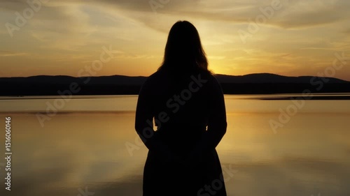Silhouette of a lonely young girl standing on the shore of a lake at sunset. The girl admires the beauty of nature. Human privacy.