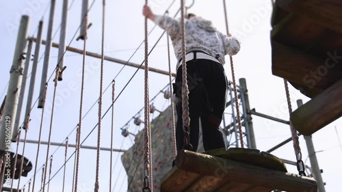 View of high ropes course, kids of climbing in amusement acitivity rope park, passing obstacles and zip line on heights, children teenagers in equipment gear between the trees on heights, summer day