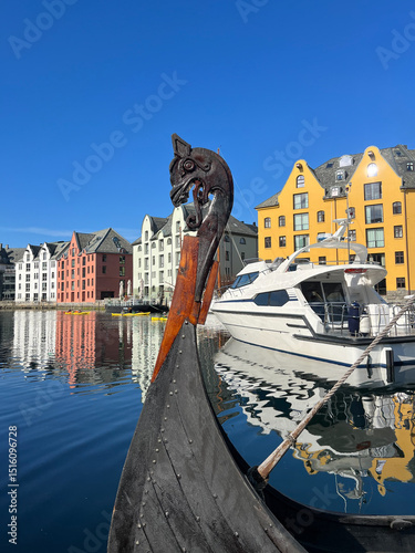 Viking figurehead on the bow of a ship anchored in the Brosundet Canal, with luxury boats and colorful Art Nouveau buildings in Ålesund, Norway, with a blue sky on a sunny day
