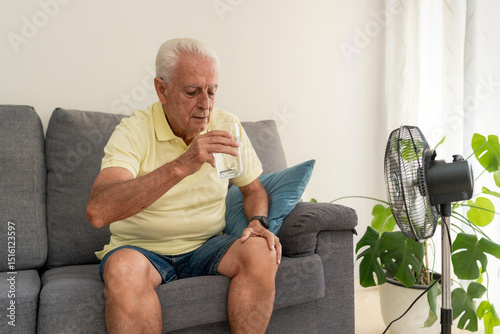 Senior man drinking water on sofa with electric fan cooling him down during summer heatwave at home