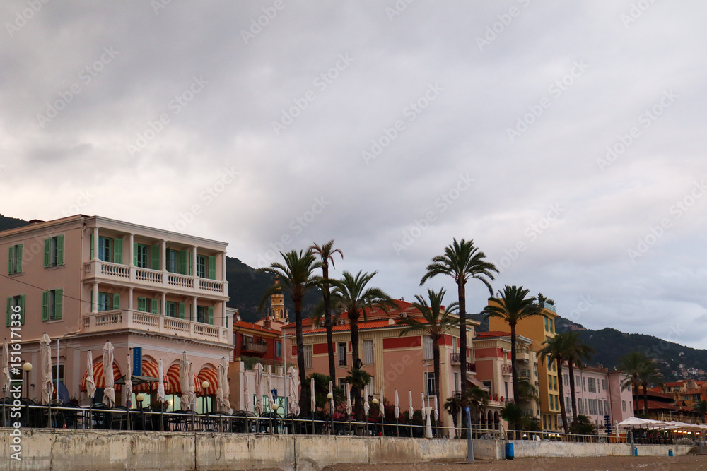 Obraz premium The pastel-coloured skyline of Menton glows at sunset, with charming rooftops and coastal silhouettes. A golden hour view of this French Riviera gem.