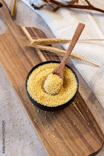 Uncooked couscous grain in wooden spoon in black bowl full of couscous and on wooden cutting board
