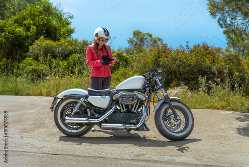 A beautiful young woman wearing a red jacket and open-face helmet putting on her gloves next to her customized Harley-Davidson motorcycle on a scenic coastal road.