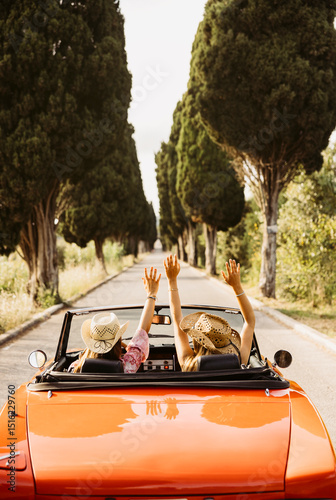 Foto Two young women raising arms while driving orange convertible car on a country r