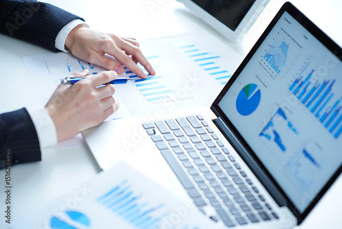 A business professional analyzing financial data charts and graphs on a laptop and paper in office