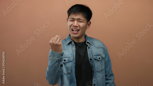 Young man expressing skepticism with hand gestures against an isolated brown wall background, conveying uncertainty and contemplation while wearing a casual denim jacket.