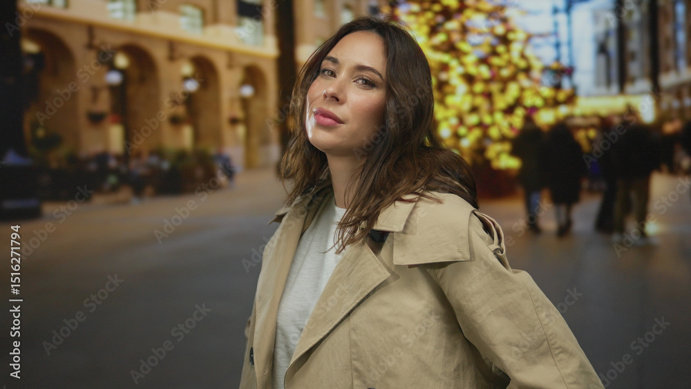 Fototapeta premium Woman in trenchcoat stands confidently on city street adorned with festive lights, radiating a warm expression in an urban setting during the holiday season.
