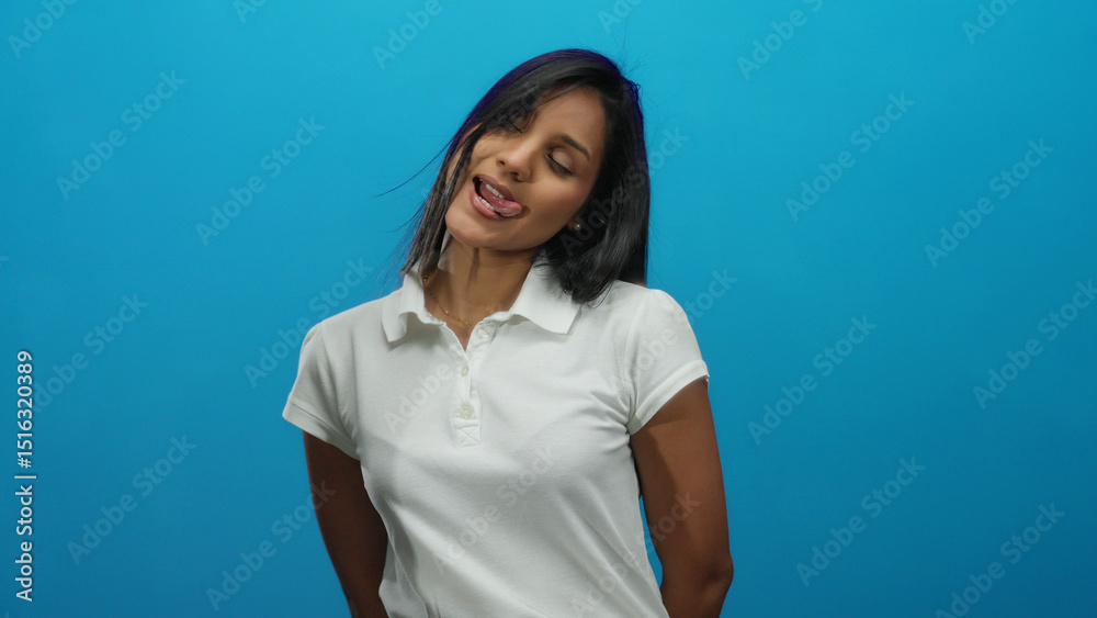 Woman smiling playfully against a vibrant blue background, showcasing her youthful energy and confident expression in a casual white shirt with long black hair. Woman smiling playfully against a vibrant blue background, showcasing her youthful energy and confident expression in a casual white shirt with long black hair.