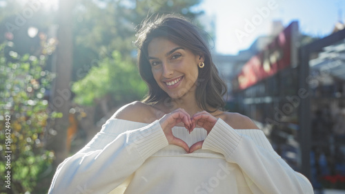 Wallpaper Mural Woman smiling in city park with sunlight highlighting her features, wearing off-shoulder sweater, making heart shape with hands, displaying joy and beauty outdoors. Torontodigital.ca
