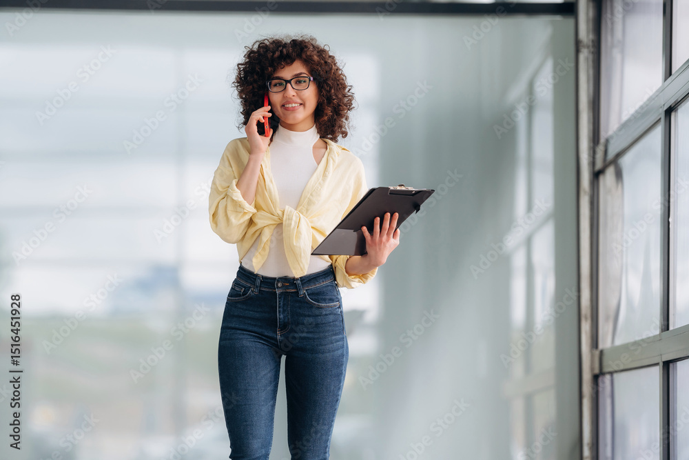 Fototapeta premium Talking by phone. Beautiful woman is standing indoors against blurred wall