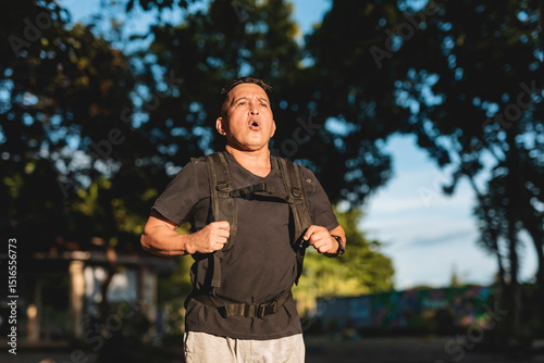 Middle aged Southeast Asian man runs through park wearing weighted backpack, regulating his breathing during an intense rucking workout. VO2 max run. photo