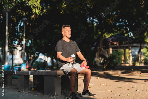 A middle aged asian man resting on a bench at the park during a hot afternoon, holding a water bottle and towel.Thinking and pondering over what to do.
