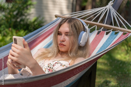 Caucasian girl 20 years old with headphones resting in a hammock and reading news on her phone