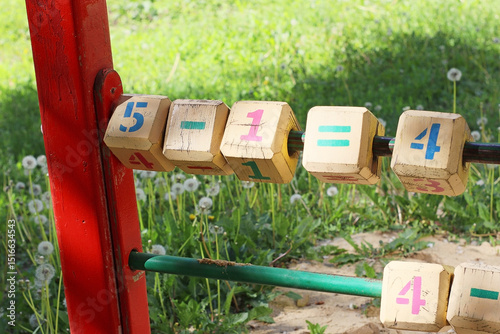Closeup of wooden educational blocks with numbers and math symbols on a playground. Outdoor learning toy for children. Sunny day with grass and dandelions in the background.