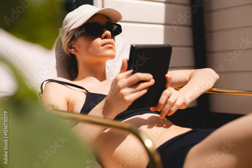 Tableau sur toile Blonde short hair girl in sunglasses, cap and swimsuit reads an e-book on a backdoor house