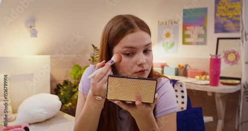 A girl does her makeup while sitting on the bed. She puts blush on her face with a brush.