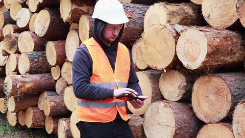 A worker in a safety vest and helmet is focused on a tablet in front of stacked logs at a lumber yard. He appears to be managing tasks and monitoring inventory in a forested setting