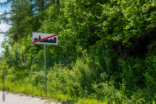 Polish traffic sign indicating the end of a built-up area, located by a forested roadside with sidewalk and greenery, under sunny weather.