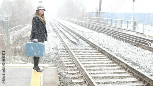 Attractive young woman waiting for a train at a train station stop in winter with falling snow, wearing vintage clothing and holding an old blue suitcase.