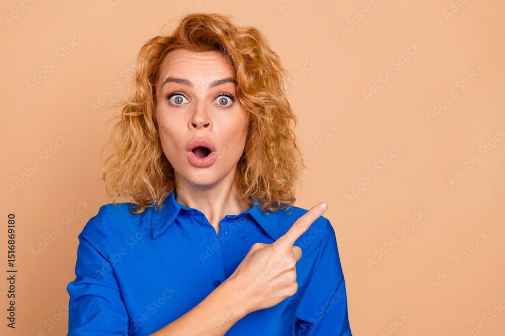 A surprised young woman in a blue shirt pointing to the side against a beige background expressing astonishment A surprised young woman in a blue shirt pointing to the side against a beige background expressing astonishment