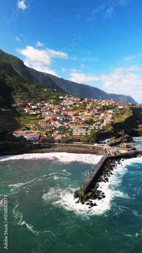 Cinematic aerial footage of a small coastal town perched on dramatic rocky cliffs along the Atlantic Ocean on Madeira Island, Portugal.