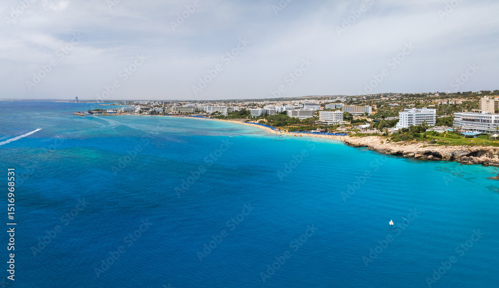 Fototapeta premium Aerial view of Ayia Napa, Cyprus, showing turquoise waters, sandy beaches, luxury resorts, white buildings, rocky outcrops, and green vegetation.