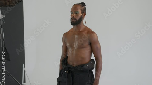 Muscular shirtless African American gay man with makeup posing for professional photographer in studio against white backdrop. Photographer capturing LGBTQ individual