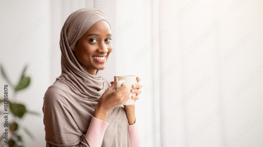Black Muslim woman in traditional hijab drinking hot aromatic coffee near window at home, having peaceful weekend. Portrait of beautiful young lady in Arabic headscarf having warming beverage indoors Black Muslim woman in traditional hijab drinking hot aromatic coffee near window at home, having peaceful weekend. Portrait of beautiful young lady in Arabic headscarf having warming beverage indoors
