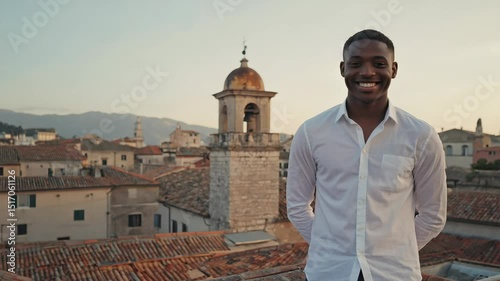 Wallpaper Mural Young, cheerful businessman smiling while standing on a rooftop, overlooking a vibrant cityscape with a stunning bell tower at sunset, capturing a moment of professional success and happiness Torontodigital.ca