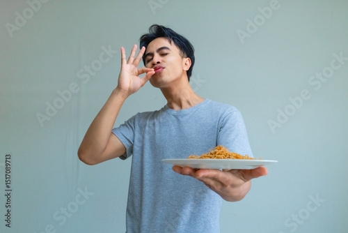 Young Asian man enjoying and savoring delicious noodles with a happy, satisfied expression, licking his lips as he eats, isolated on a clear background