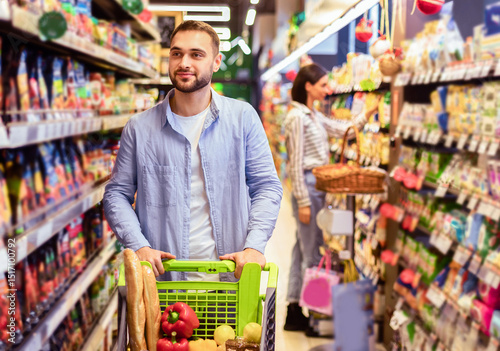 Wallpaper Mural Everyday Shopping. Portrait Of Young Bearded Guy Walking With Trolley Cart Between The Aisles In Wholesale Store. Casual Man Buying Healthy Food In Shop, Looking At Shelves, Blurred Background Torontodigital.ca