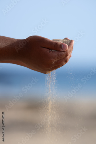A hand is holding a handful of sand and it is pouring out of the hand. The sand is falling onto the beach, creating a sense of movement and energy. The image conveys a feeling of relaxation