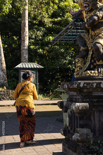 Balinese woman in traditional dress at a temple with religious sculptures in the afternoon with heavy shadows in Indonesia.