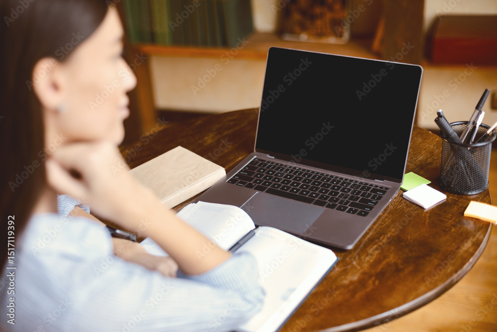 Fototapeta premium E-learning Concept. Over the shoulder view of woman sitting at desk with open notebook, using laptop with black empty screen for mockup template, watching webinar, studying online at home