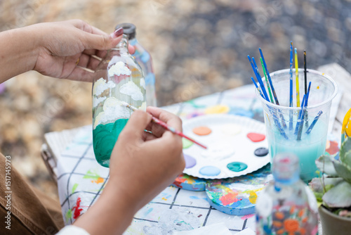 Person creates sustainable and ecofriendly craft art by painting clouds and sky on glass bottle using fine brushes and colors, expressing creativity and environmental care