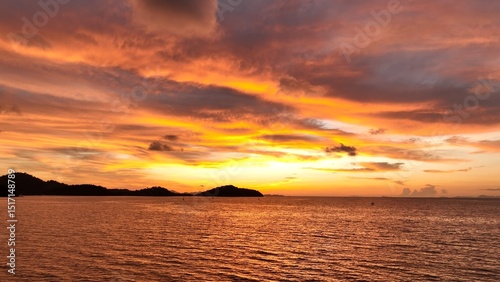 A serene beach sunset, where the sky is ablaze with colors of orange and pink, as the sun sets behind distant hills, casting a warm glow across the peaceful ocean and inviting relaxation. Thailand.
