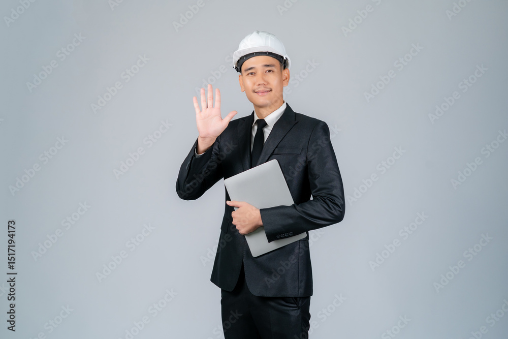 Young Asian engineer or architect raising his hand in greeting and holding a laptop in his hand on a background. Young Asian engineer or architect raising his hand in greeting and holding a laptop in his hand on a background.