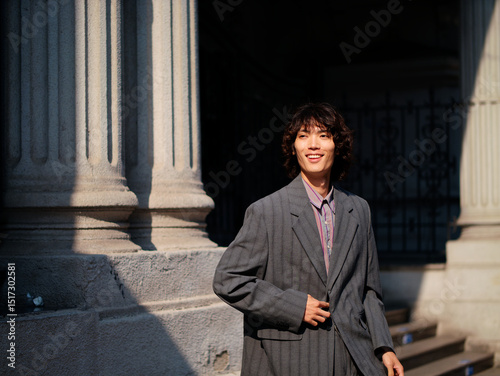 Portrait of handsome Chinese young man wearing gray suit posing in the street, young guy with black curly hair with urban background. Male fashion, cool Asian young man lifestyle.
