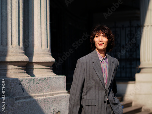 Portrait of handsome Chinese young man wearing gray suit posing in the street, young guy with black curly hair with urban background. Male fashion, cool Asian young man lifestyle.