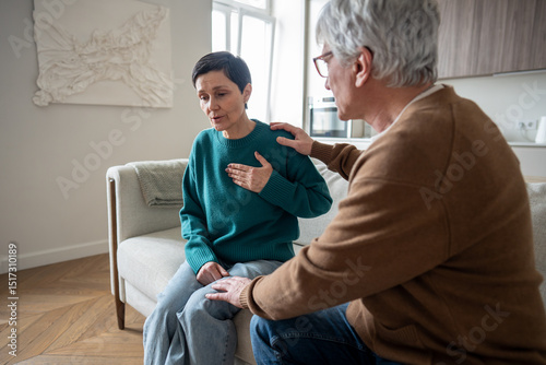 Scared elderly woman figure gasps clutches heart with growing panic as worried husband watches helplessly trying to calm. Sudden chest pressure, breathing difficulty, heart shock, unclear what to do