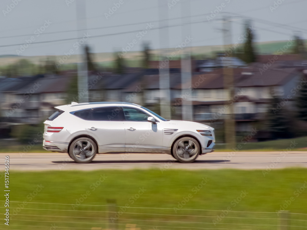 Fototapeta premium Calgary, Alberta, Canada. Jun 2, 2025. A sleek white Genesis GV80 moves gracefully on an urban road, its luxurious design highlighted by motion blur against a suburban residential backdrop.