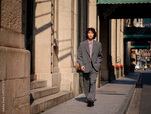 Portrait of handsome Chinese young man wearing gray suit walking in the street, young guy with black curly hair with urban background. Male fashion, cool Asian young man lifestyle.