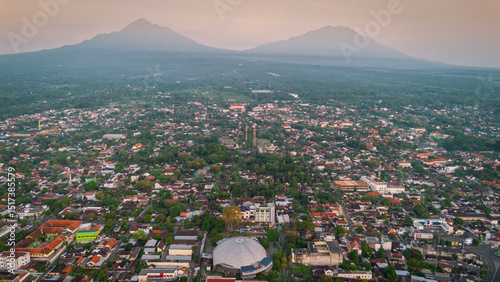 An aerial perspective captures an Indonesian city nestled between two majestic mountains, showcasing a dense urban sprawl with diverse architecture. The image is taken from a high angle.