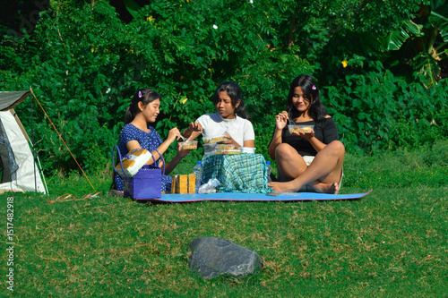Sitting on a picnic mat in front of the tent, the teenage girls resume their meal while enjoying the relaxing natural surroundings. photo