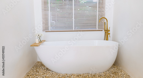 An elegant white freestanding bathtub rests on pebbles in a minimalist bathroom. Complemented by a golden faucet and a wooden tray with flowers and a candle.