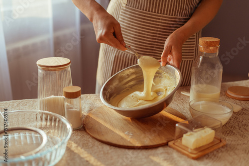 Photos A person wearing a striped apron stirs thick batter in a metal bowl with a spoon, surrounded by baking ingredients on a table