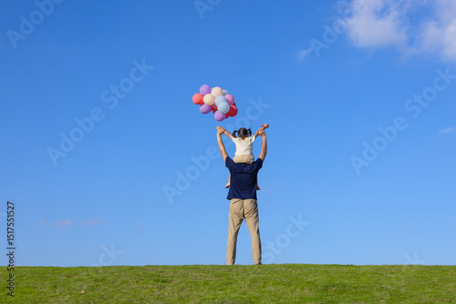 A little girl playing with balloons on the grass is sitting on her father's shoulders