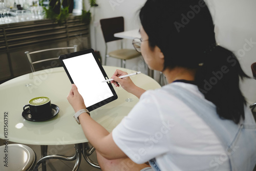 Woman using digital tablet with stylus pen at coffee shop, blank screen ready for design or note-taking, with matcha latte on round glass table.