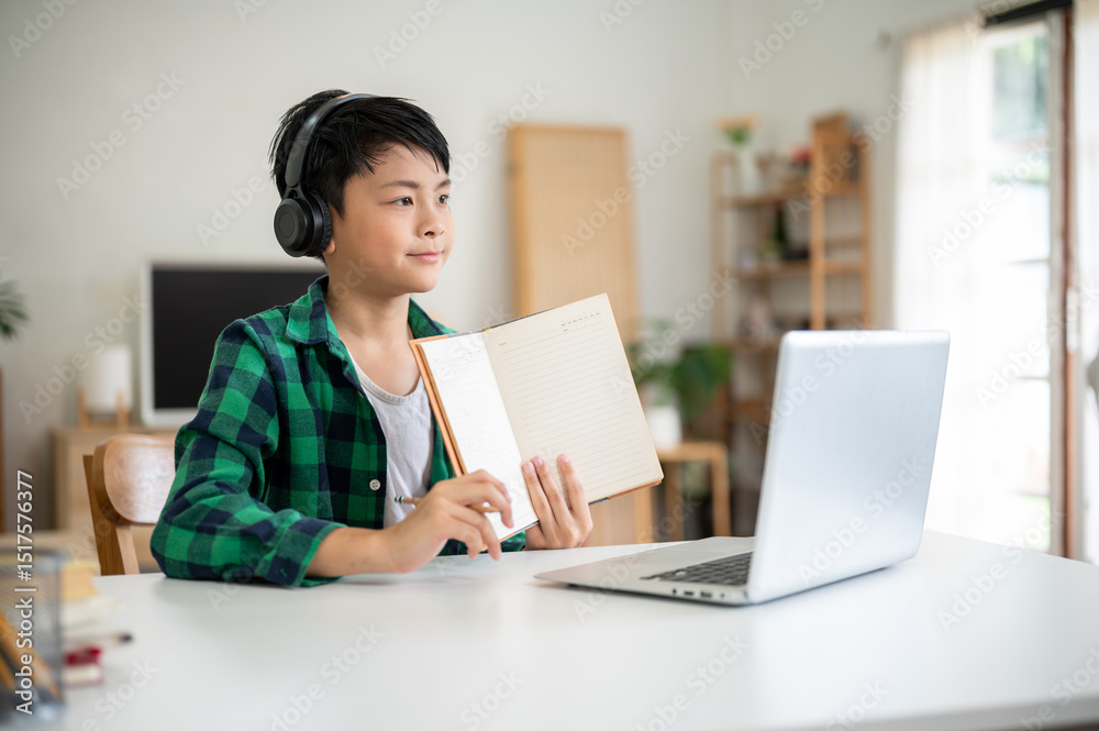 Asian kid elementary school boy wearing headphones holding a notebook in front of a laptop on table. Asian kid elementary school boy wearing headphones holding a notebook in front of a laptop on table.